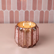 A lit Sweet Grace Collection Candle #021 by Bridgewater Candle Company with two flames glows in a faceted rose gold mercury glass holder of soy-blend wax, resting on a light pink surface against a pink and white tiled background.