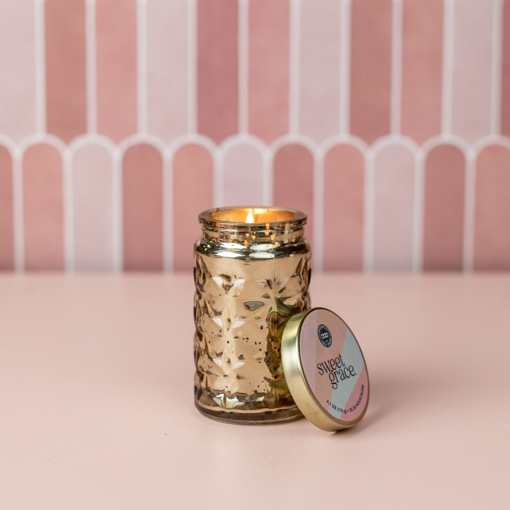A Bridgewater Candle Company Sweet Grace Collection Candle #022 in a textured gold glass jar sits on a pink surface with its lid beside it, showcasing its premium scent against a geometric pink and white tile backdrop.