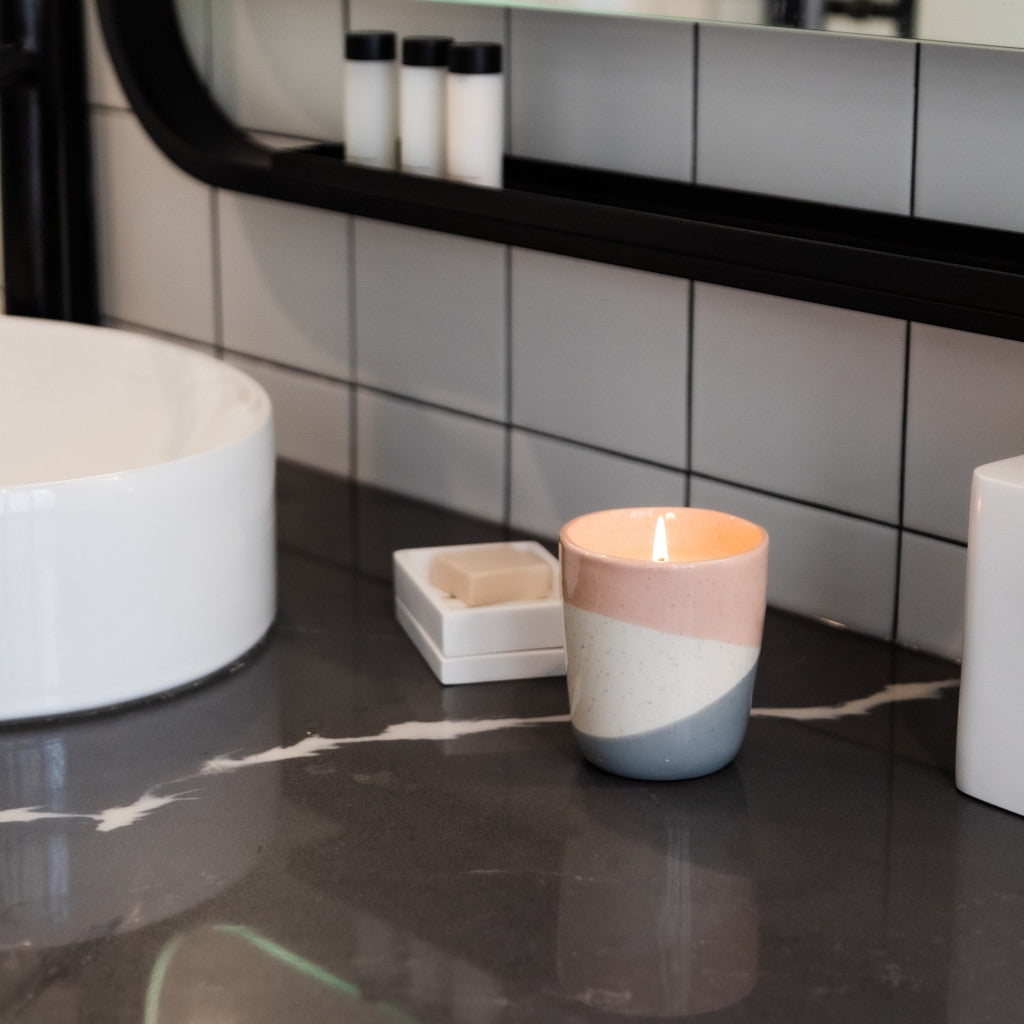 A Bridgewater Candle Company Sweet Grace Collection Candle #041 in a pastel holder sits on a dark marble bathroom counter by a white soap dish, filling the space with its best-selling fragrance, modern sink and tiled wall in view.