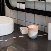 A Bridgewater Candle Company Sweet Grace Collection Candle #041 in a pastel holder sits on a dark marble bathroom counter by a white soap dish, filling the space with its best-selling fragrance, modern sink and tiled wall in view.