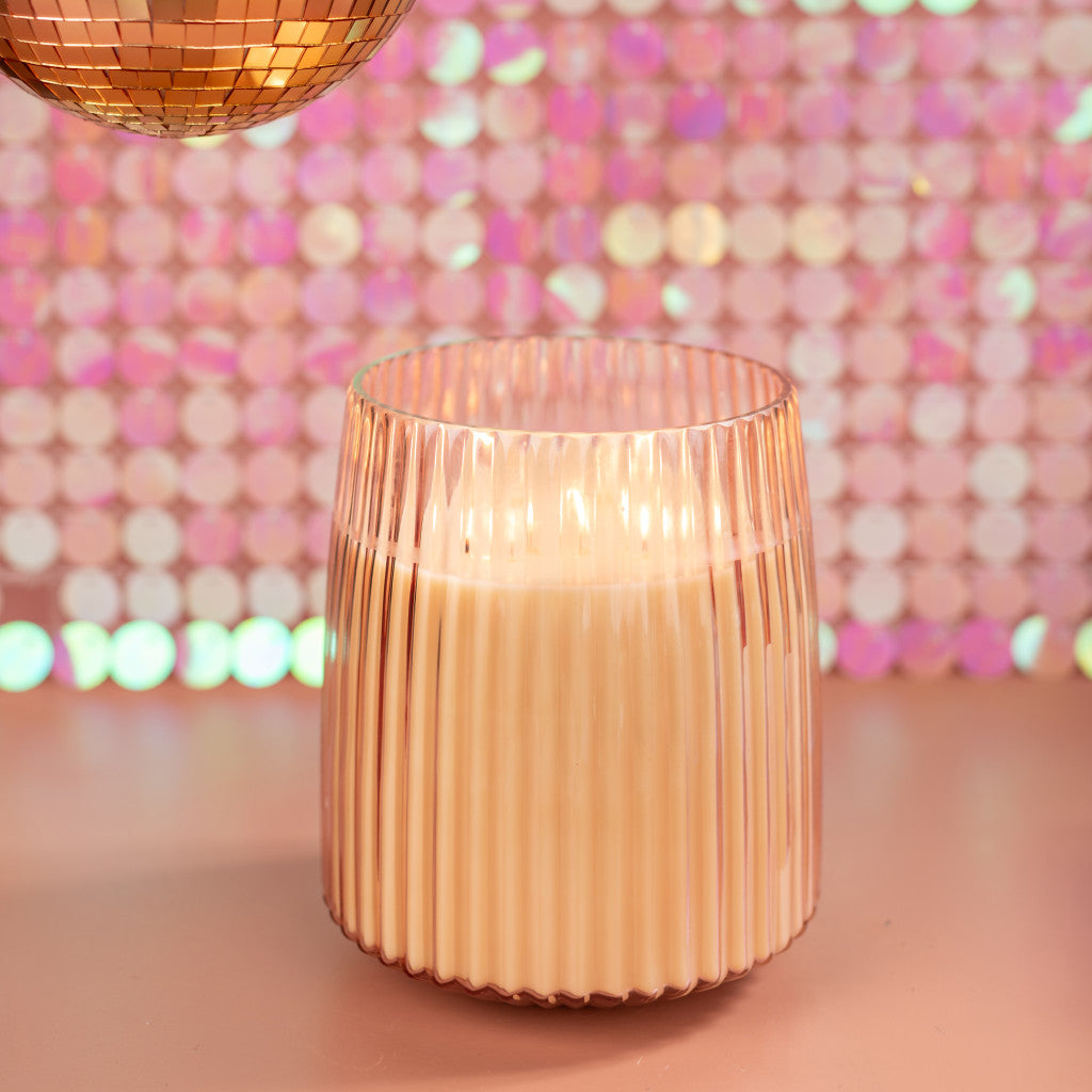 The Sweet Grace Collection Candle #043 by Bridgewater Candle Company, made from soy-blend wax, glows in a ribbed glass holder on a pink surface with a sparkly pink sequin backdrop and a gold disco ball above.