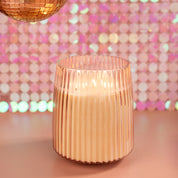 The Sweet Grace Collection Candle #043 by Bridgewater Candle Company, made from soy-blend wax, glows in a ribbed glass holder on a pink surface with a sparkly pink sequin backdrop and a gold disco ball above.