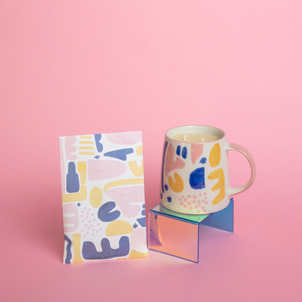 A mug with abstract yellow, pink, and blue shapes sits on a small iridescent stand next to a matching pouch and the Sweet Grace Collection Candle #047 by Bridgewater Candle Company, all arranged against a pink background.