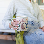 A person in a striped shirt and jeans sits on a green chair, holding the Bridgewater Candle Company Sweet Grace Collection Candle #048.