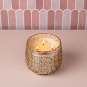 A lit Sweet Grace Collection Candle #051 by Bridgewater Candle Company, featuring soy-blend wax in a textured metallic gold holder, sits on a pale pink surface with a pink and white arched tile background.
