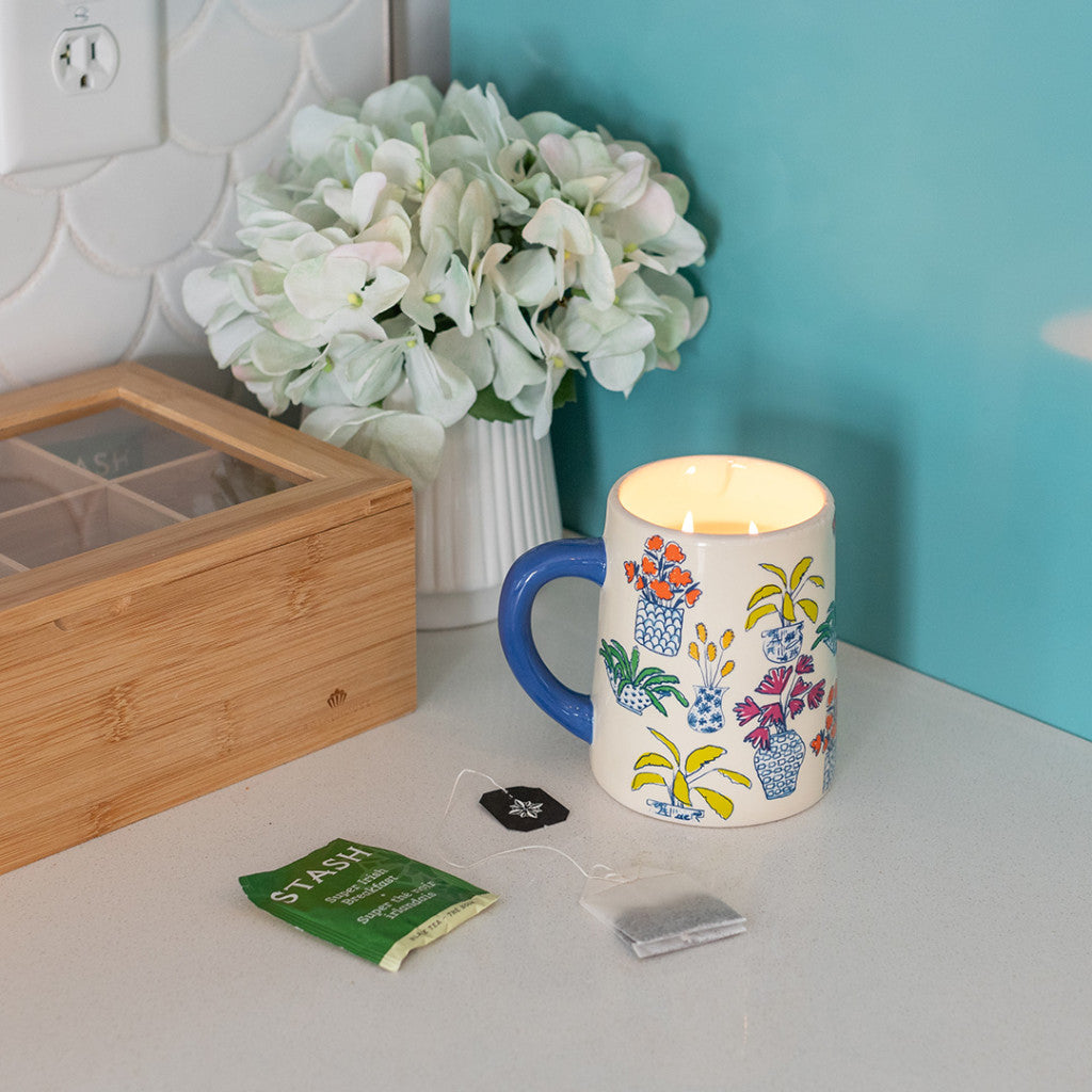 A floral mug with a lit Bridgewater Candle Company Sweet Grace Collection Candle #054 sits on a counter beside a wooden tea box, an unopened green tea packet, a used tea bag, and a white vase of white hydrangeas—a plant mom’s dream.