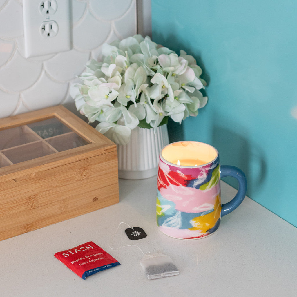 A Sweet Grace Collection Candle #055 by Bridgewater Candle Company sits on a white countertop beside a colorful mug, small white vase of flowers, wooden tea box, and an unused Stash tea bag and packet.
