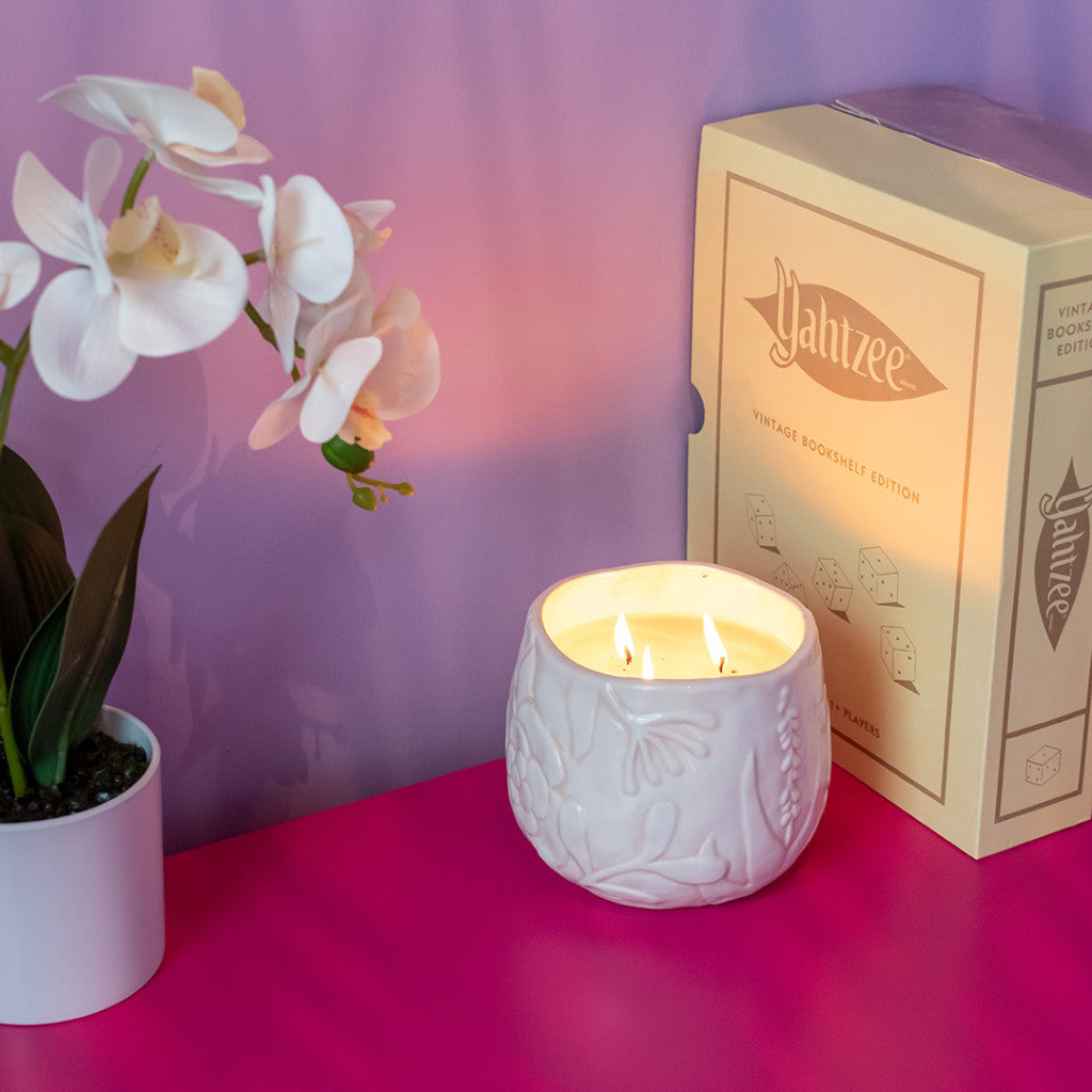 A white orchid plant, a Bridgewater Candle Company Sweet Grace Collection Candle #057, and a yellow Yahtzee Vintage Bookshelf Edition box rest on a bright pink surface against a purple wall.