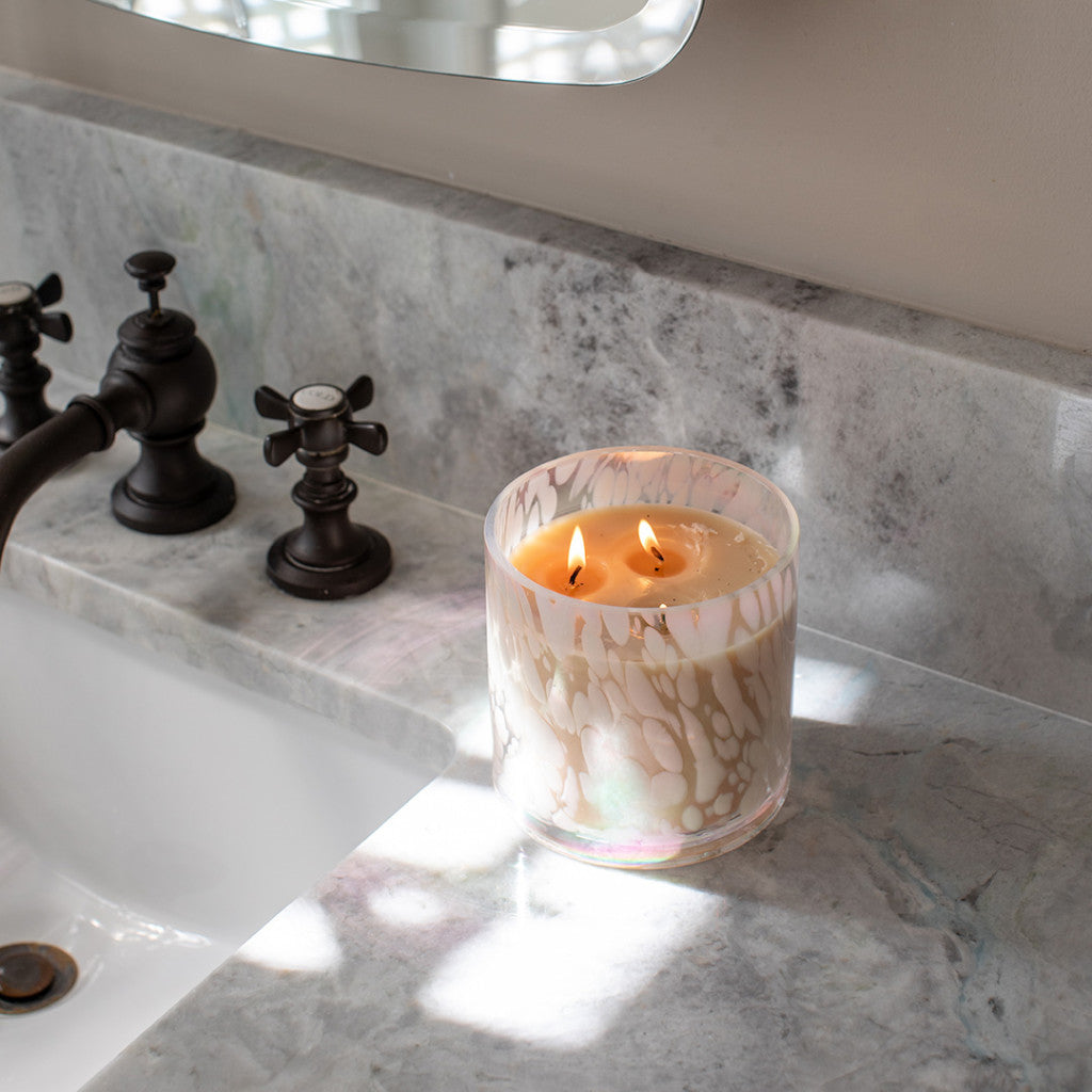 A marbled glass Sweet Grace Collection Candle #058 by Bridgewater Candle Company, featuring two flames and soy-blend wax, burns on a gray marble bathroom countertop beside a sink and black faucet.