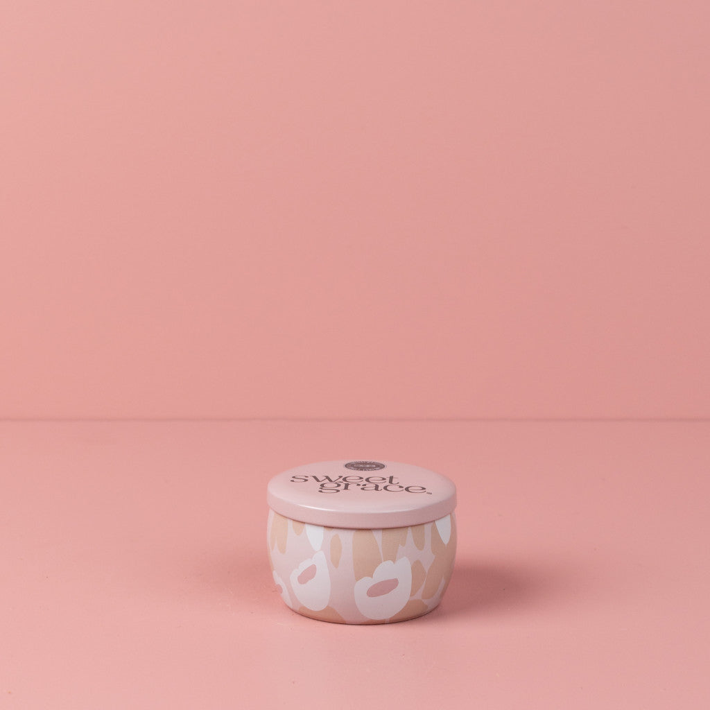 The Bridgewater Candle Company Sweet Grace Collection Candle #059 is a small, round travel candle with a light pink lid and white abstract design. Crafted from premium soy-blend wax, it offers soothing aromas and sits on a matching pink surface.