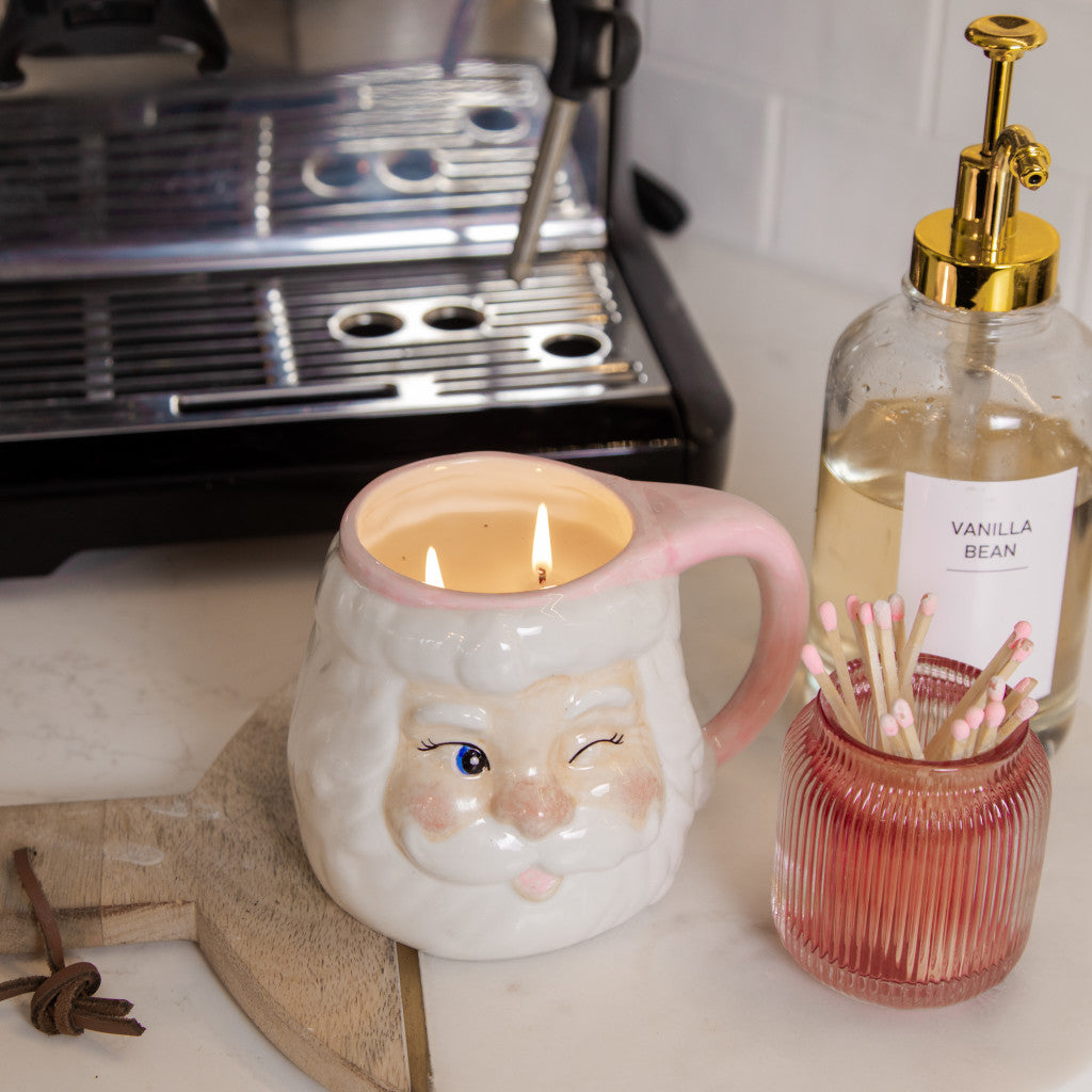 A reusable Sweet Grace Santa Mug Candle by Bridgewater Candle Company, shaped like a winking Santa, glows warmly alongside a glass Vanilla Bean pump bottle, pink match jar, and an espresso machine—creating a festive holiday candle setting.