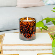 A lit Bridgewater Candle Company Sweet Grace Collection Candle #064, in a brown tortoise glass holder, sits on a white and wood box with a green plant and light gray sofa in the background. The soy-blend wax creates an inviting atmosphere.