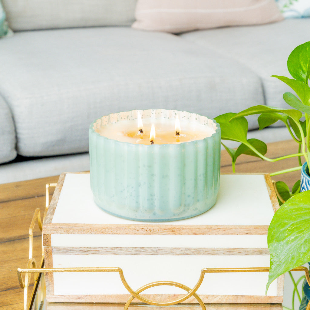 A lit Sweet Grace Collection Candle #065 by Bridgewater Candle Company in an antique green ribbed vessel sits on a stack of books on a wooden coffee table, with a nearby green plant and a light-colored sofa in the background.