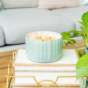 A lit Sweet Grace Collection Candle #065 by Bridgewater Candle Company in an antique green ribbed vessel sits on a stack of books on a wooden coffee table, with a nearby green plant and a light-colored sofa in the background.