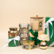 A group of elegant green and cream gift boxes with ribbons, an Afternoon Retreat Mercury Flower Diffuser by Bridgewater Candle Company, a jar of gold glitter, and a white artificial flower in a gold vase on a beige background.