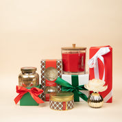 A festive display featuring the Bridgewater Candle Company Christmas Bliss Holiday 3-Wick Tin Candle, a gold ornament vase with a white flower, and gifts wrapped in red, white, and green ribbons—all set against a beige background.