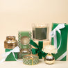 A selection of green and gold holiday gifts, featuring Bridgewater Candle Company's Festive Frasier Holiday Mercury Flower Diffuser and green-wrapped presents with white ribbons, set against a light beige background.