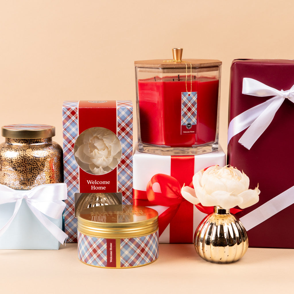 A selection of gifts, including plaid tins, a large red candle, and Bridgewater Candle Company’s Welcome Home Mercury Flower Diffuser with a gold-silver finish—all beautifully arranged on beige for an elegant home fragrance touch.