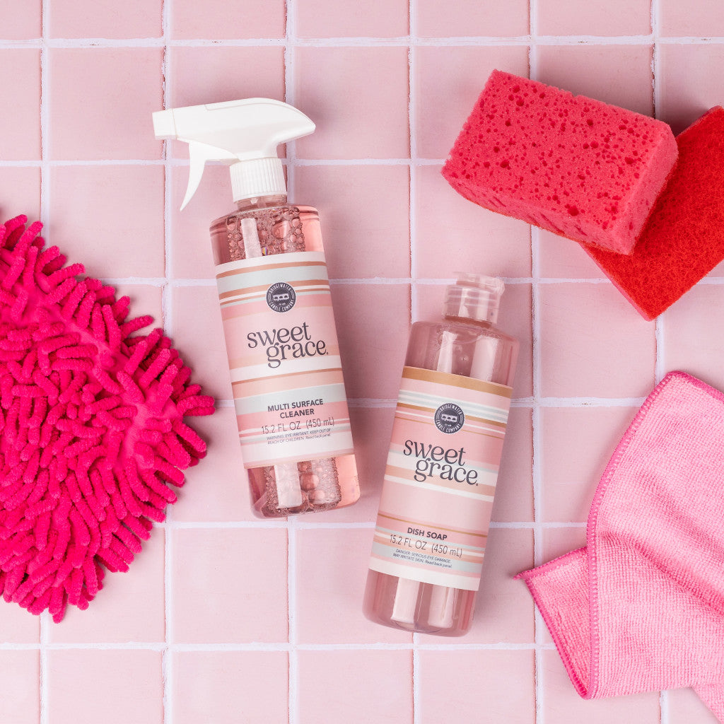 Two pink cleaning cloths, a red sponge, and two bottles of Bridgewater Candle Companys Sweet Grace Multi-Surface Cleaner add fragrance to the pink-tiled surface.