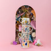 Four colorful patterned mugs are stacked by an arched window with dried flowers and ornaments, next to the Sweet Grace Collection Candle #047 from Bridgewater Candle Company, all set against a pastel pink background.
