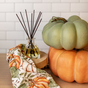 The Bridgewater Candle Company Harvest Pumpkin Reed Diffuser fills your home with a warm pumpkin fragrance, as black reeds rest in a glass jar on a wooden stand beside pumpkins and autumn accents against white tiled walls.
