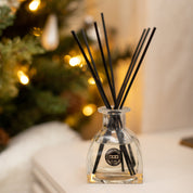 A Bridgewater Candle Company Festive Frasier Reed Diffuser with black fiber reeds rests on a light surface, while a softly glowing, decorated Christmas tree in the background fills the room with its cozy holiday fragrance.