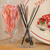 A glass jar of Bridgewater Candle Company’s Christmas Bliss Reed Diffuser fills the air with festive scent, sitting beside a candy cane jar and a plate with a red truck and Happy—a charming home décor accent.
