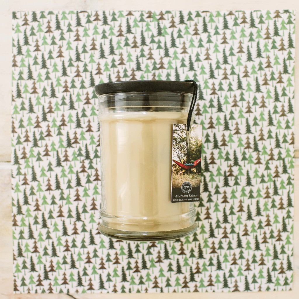 A cream-colored Bridgewater Candle Company Afternoon Retreat 8oz Jar Candle with a black lid sits on pine-patterned paper. Its label shows a hammock between trees, capturing the calm of an afternoon escape.