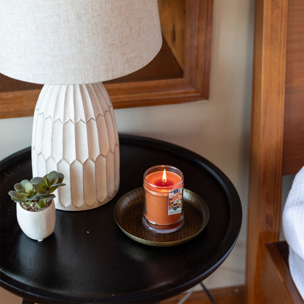 A lit Harvest Pumpkin 8oz Jar Candle by Bridgewater Candle Company sits on a round tray atop a black bedside table, beside a potted succulent and textured white lamp, with part of a bed and wooden headboard visible.
