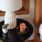 A lit Harvest Pumpkin 8oz Jar Candle by Bridgewater Candle Company sits on a round tray atop a black bedside table, beside a potted succulent and textured white lamp, with part of a bed and wooden headboard visible.