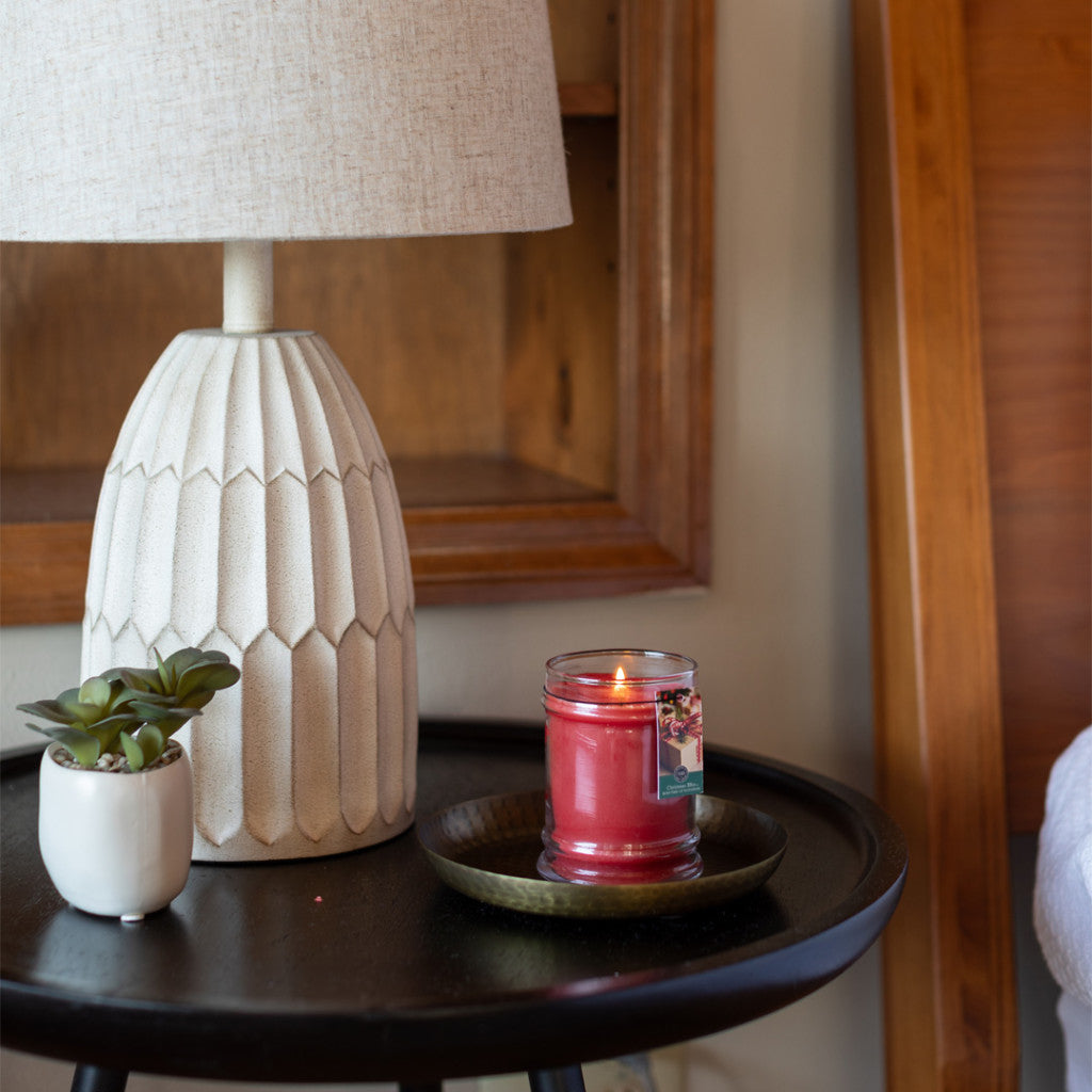 A round black side table holds a white textured lamp, a small potted succulent, and a lit Christmas Bliss 8oz Holiday Jar Candle by Bridgewater Candle Company on a plate, beside a wooden bed frame.