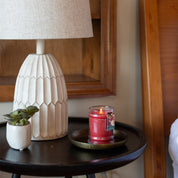 A round black side table holds a white textured lamp, a small potted succulent, and a lit Christmas Bliss 8oz Holiday Jar Candle by Bridgewater Candle Company on a plate, beside a wooden bed frame.