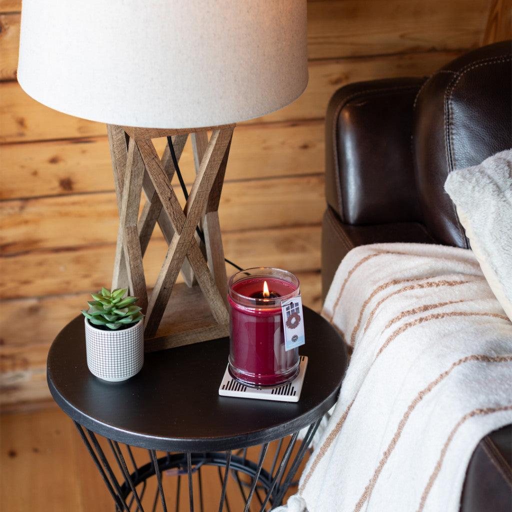A round black side table holds a small potted succulent, a Bridgewater Candle Company Welcome Home 18oz Large Jar Candle, and a wooden lamp with a white shade beside a brown leather couch draped with a striped blanket.