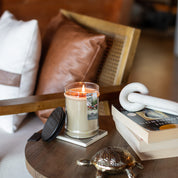 An Afternoon Retreat 18oz Large Jar Candle by Bridgewater Candle Company sits on a wooden side table beside a gold turtle, closed book, and white sculpture. A cushioned wooden chair with leather and white pillows is in the background.
