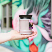 A hand holds a Bridgewater Candle Company Kiss in the Rain 18oz Large Jar Candle with a pink label, set against a vibrant mural featuring large, abstract floral designs.
