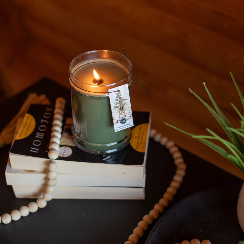 A lit Festive Frasier 18oz Large Jar Candle by Bridgewater Candle Company glows atop two stacked books, accompanied by wooden beads and a small potted plant on a dark surface, creating a warm, cozy ambiance.