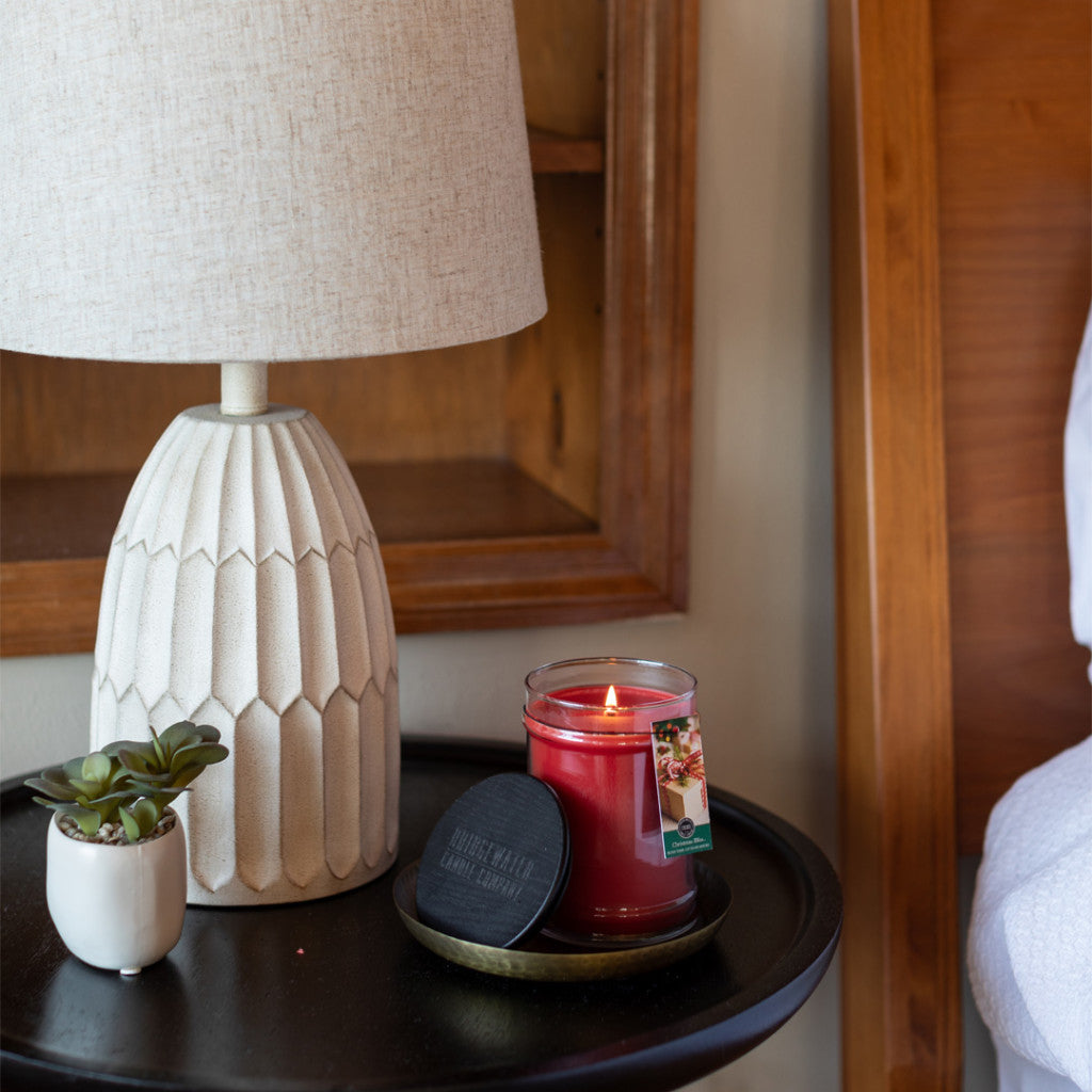 A lit Bridgewater Candle Company Christmas Bliss 18oz Large Jar sits on a round black nightstand beside a small potted succulent and a textured white ceramic lamp. The candle lid rests nearby, with a wooden headboard visible on the right.
