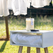 A lit Laundry Line 18oz Large Jar Candle by Bridgewater Candle Company sits on a small white table with a folded towel underneath, outdoors as fresh laundry dries in the background, filling the air with its fresh scent.