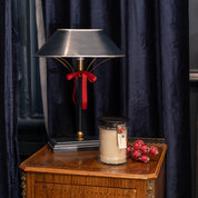 A wooden side table holds a Bridgewater Candle Company Comfort & Joy 18oz Large Jar Candle in beige, red decorative berries, and a black and silver lamp with a red ribbon, adding warmth against dark blue curtains.