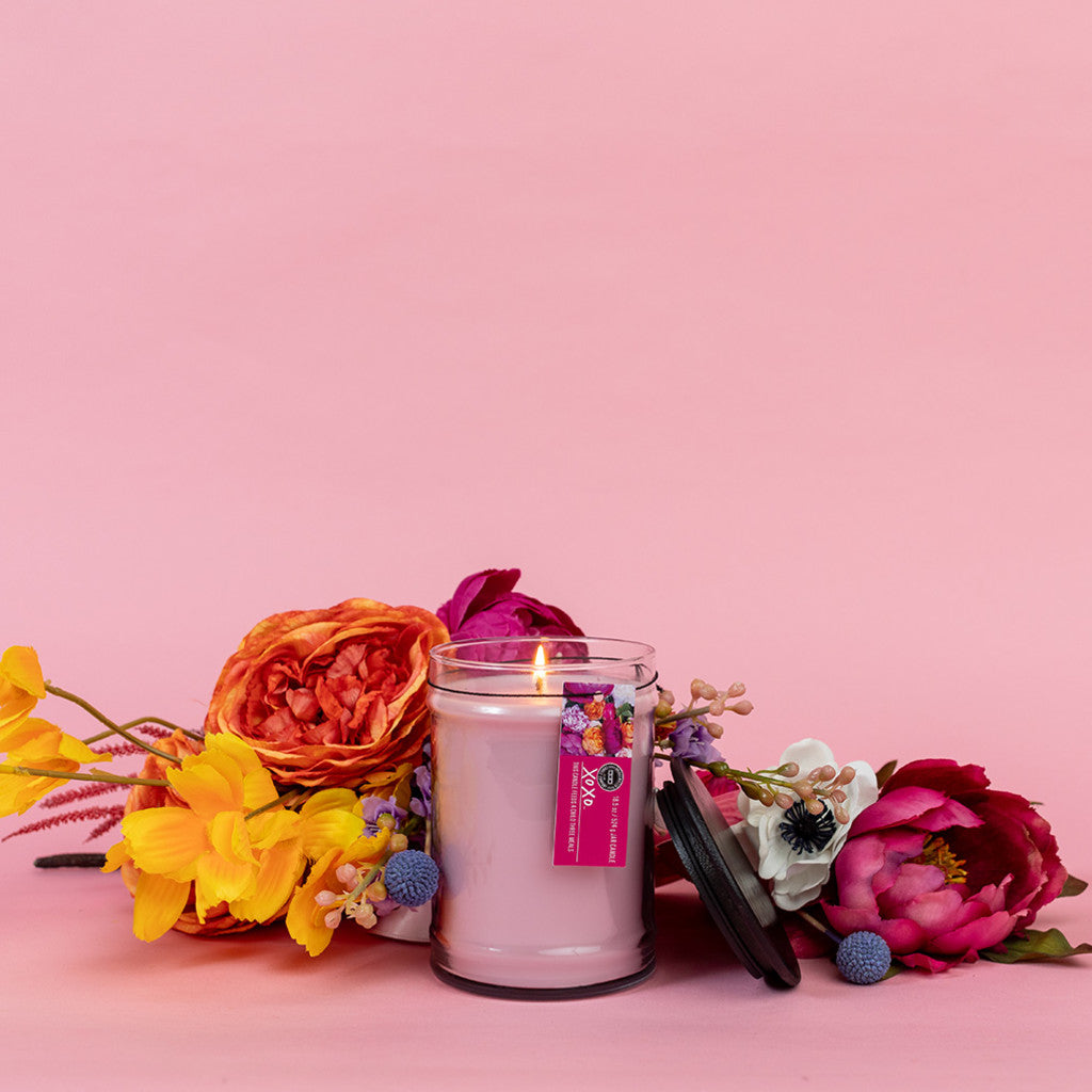 A lit XOXO 18oz Large Jar Candle by Bridgewater Candle Company with soy-blend wax sits on a pink surface, surrounded by colorful artificial flowers, with its black lid resting nearby.