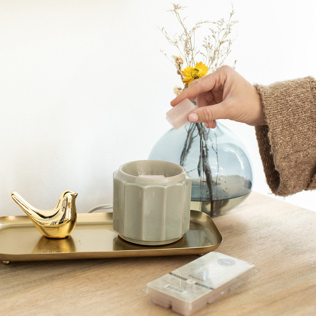 A hand places a wax melt into a light green wax warmer on a wooden table. A gold bird figurine and a clear vase with dried flowers are on a gold tray nearby.
