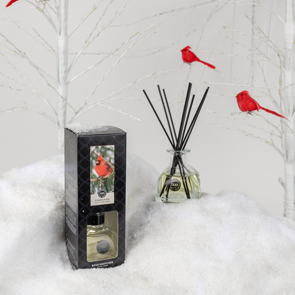A Bridgewater Candle Company Comfort & Joy Reed Diffuser with sleek black fiber reeds sits on artificial snow, surrounded by white branches and red cardinal bird accents, creating a festive home fragrance display.