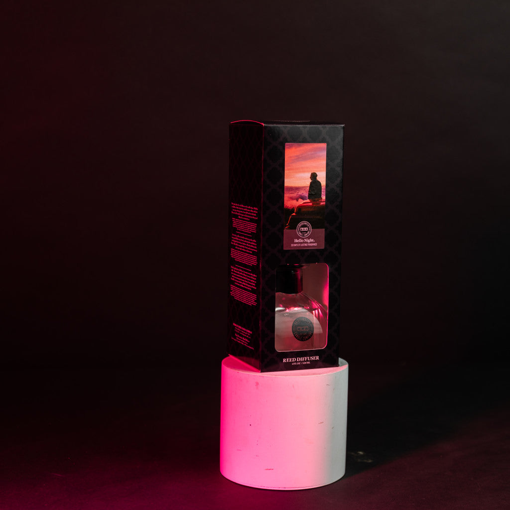 A boxed Hello Night Reed Diffuser by Bridgewater Candle Company with fiber reeds and scenic packaging is displayed on a white cylindrical pedestal, illuminated by pink and red light against a dark background.