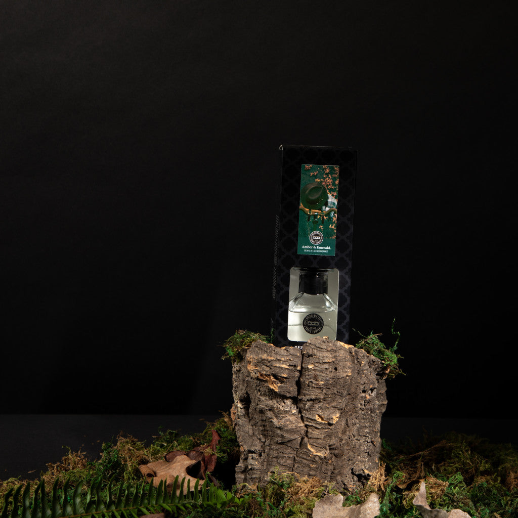 A bottle of Amber & Emerald Reed Diffuser by Bridgewater Candle Company, displayed on tree bark with moss and leaves against a dark backdrop, evokes the fresh, premium fragrance signature to Bridgewater Candle Company.