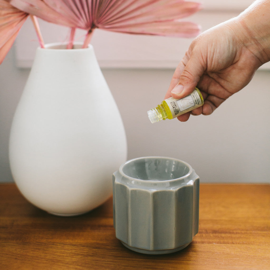A hand pours Bridgewater Candle Company’s Sweet Grace Home Fragrance Oil into a gray ceramic diffuser on a wooden table, creating flameless fragrance. A white vase with pink dried leaves is in the background.