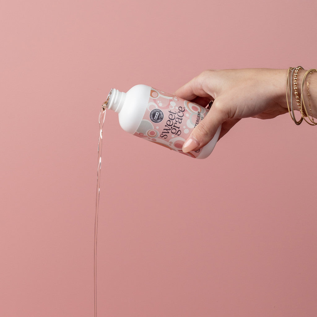 A hand wearing gold bracelets pours Bridgewater Candle Companys Sweet Grace 6oz Laundry Detergent, loved for its long-lasting, fabric-safe scent, from a bottle against a plain pink background.