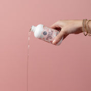 A hand wearing gold bracelets pours Bridgewater Candle Companys Sweet Grace 6oz Laundry Detergent, loved for its long-lasting, fabric-safe scent, from a bottle against a plain pink background.