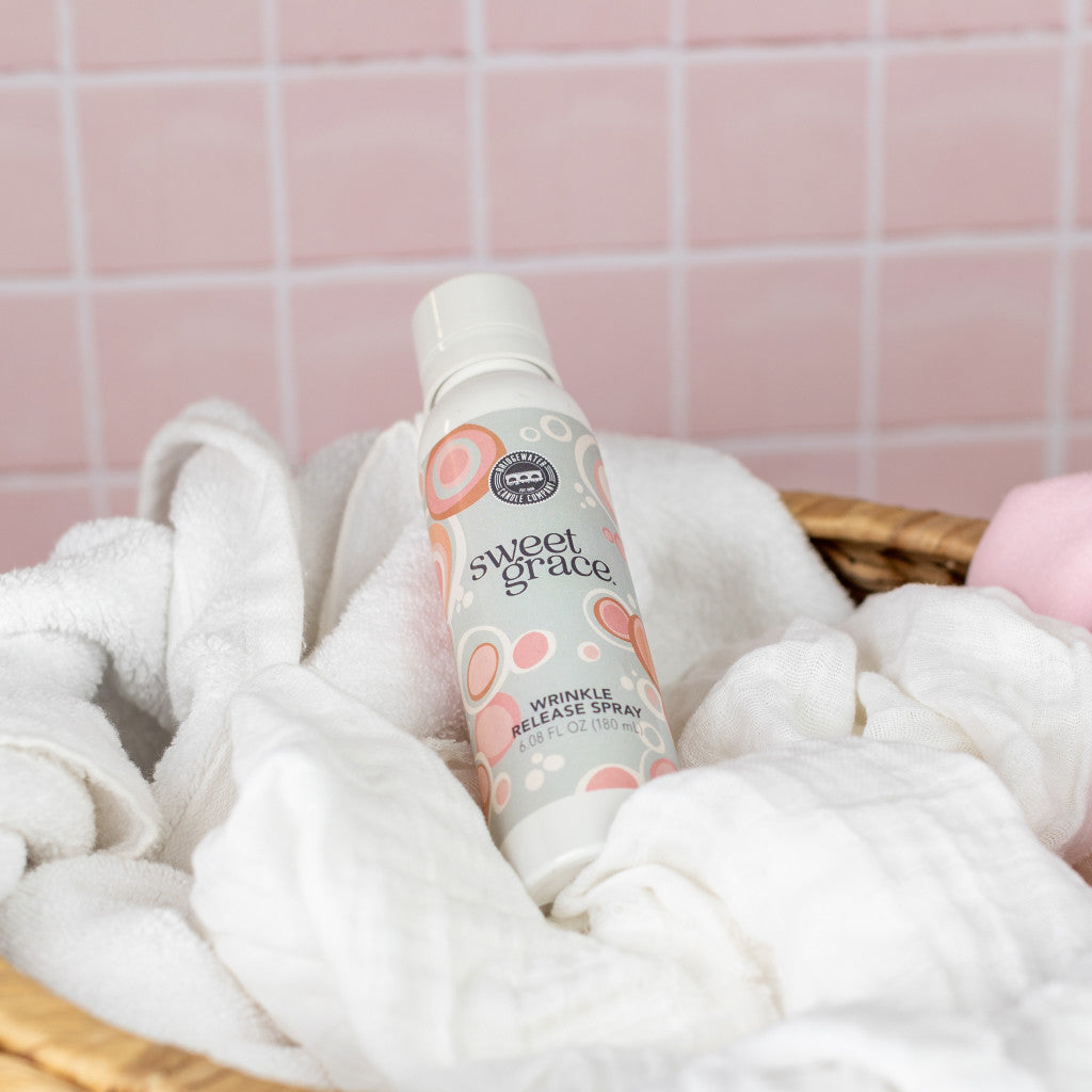 A bottle of Bridgewater Candle Company Sweet Grace Wrinkle Release Spray sits atop white towels in a woven basket against a pink tiled wall, ready to freshen up your linens with its delightful scent.