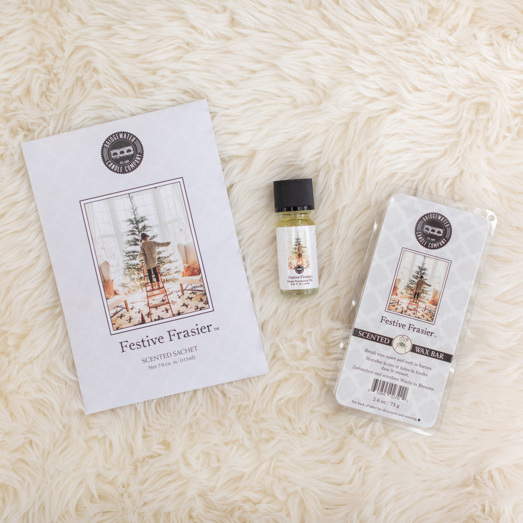 The Festive Frasier Flameless Bundle from Bridgewater Candle Company—home fragrance oil, scented sachet, and soy-blend wax bar—is displayed on a cream rug. Each comes in holiday tree-themed packaging.