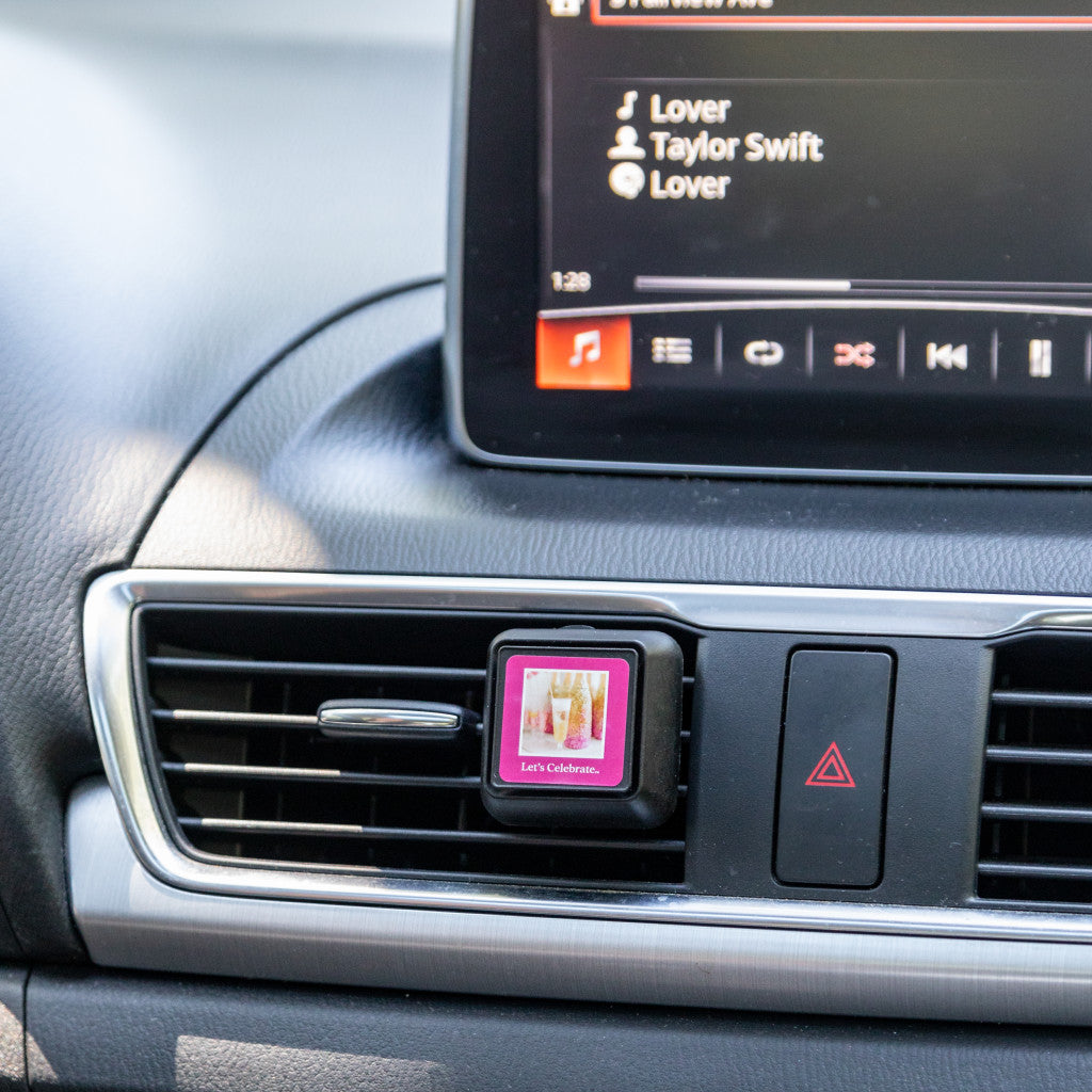 A Bridgewater Candle Company Lets Celebrate Auto Vent Clip is attached to the car dashboard, offering a floral fragrance as Lover by Taylor Swift plays on the infotainment screen.